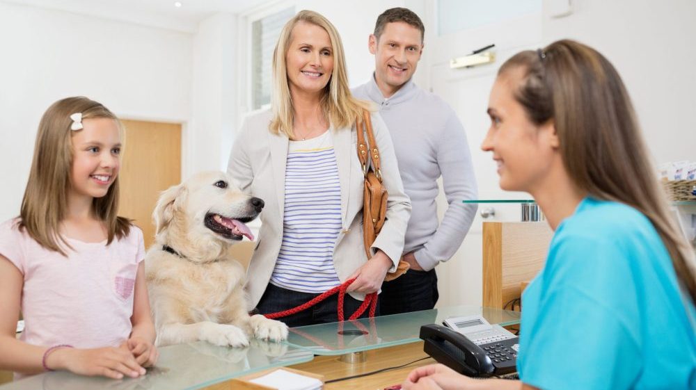 A family of a man, woman and little girl stood smiling at a veterinary receptionist with their dog