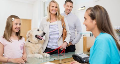 A family of a man, woman and little girl stood smiling at a veterinary receptionist with their dog